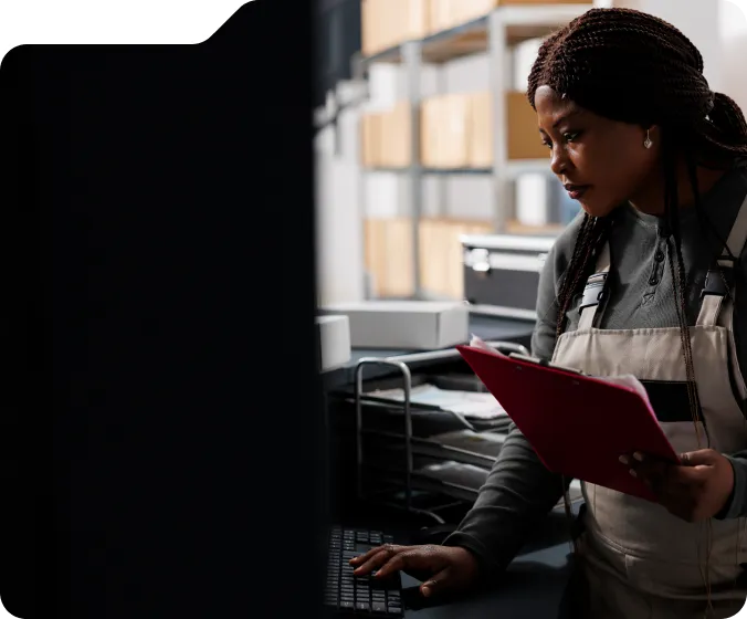 Woman in overalls holding a red clipboard typing on a keyboard in a storage room with shelves filled with boxes.