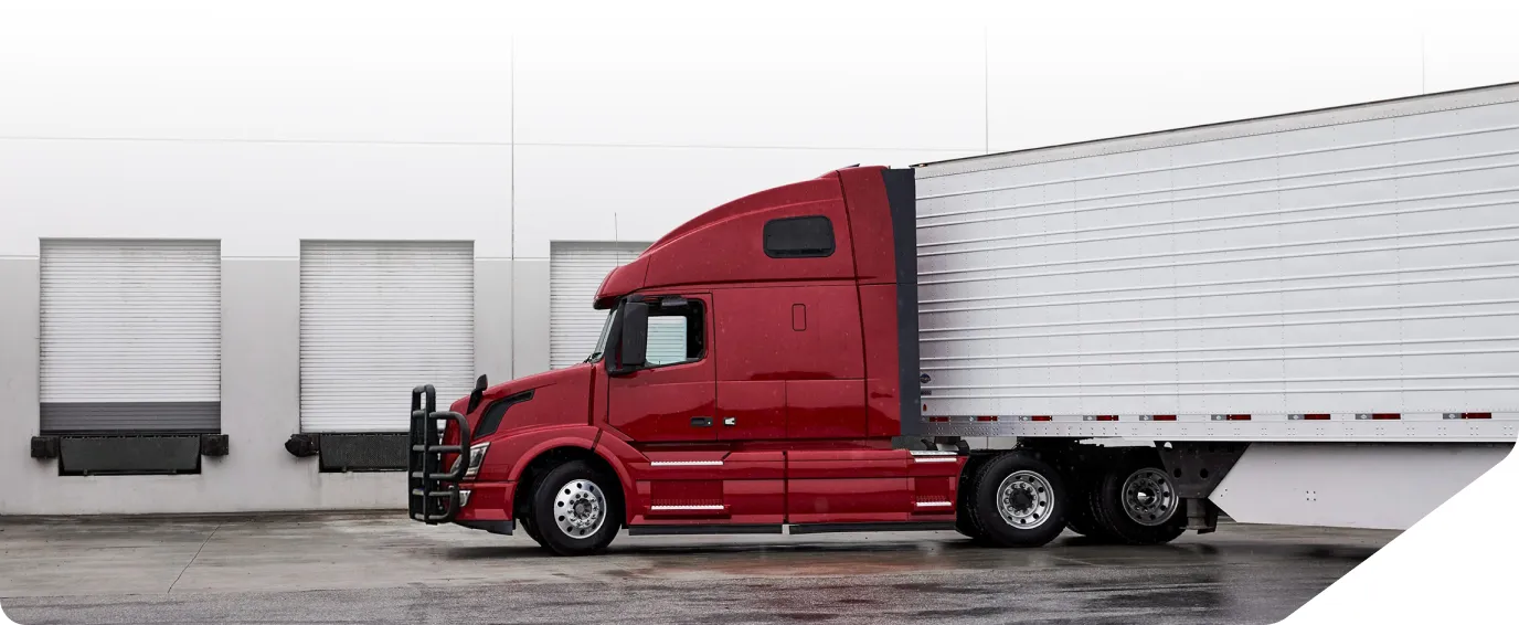Red semi-truck with white trailer parked beside a warehouse loading dock with closed roll-up doors.