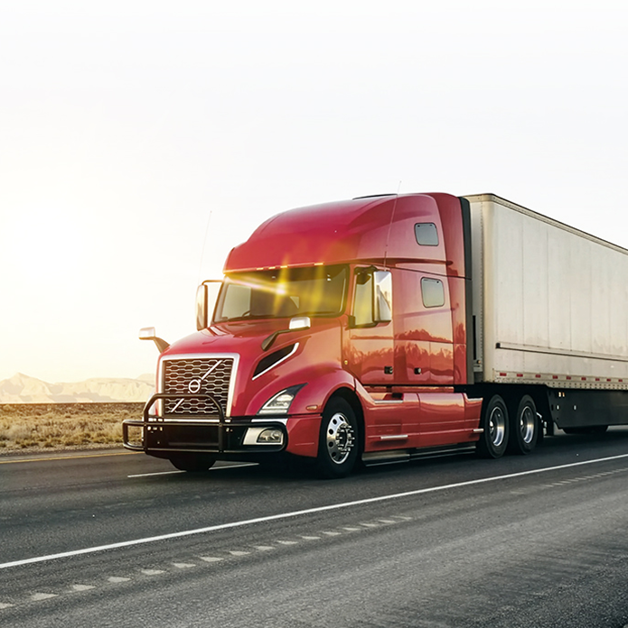 Red semi-truck with large white trailer driving on highway at sunset with desert landscape in background.