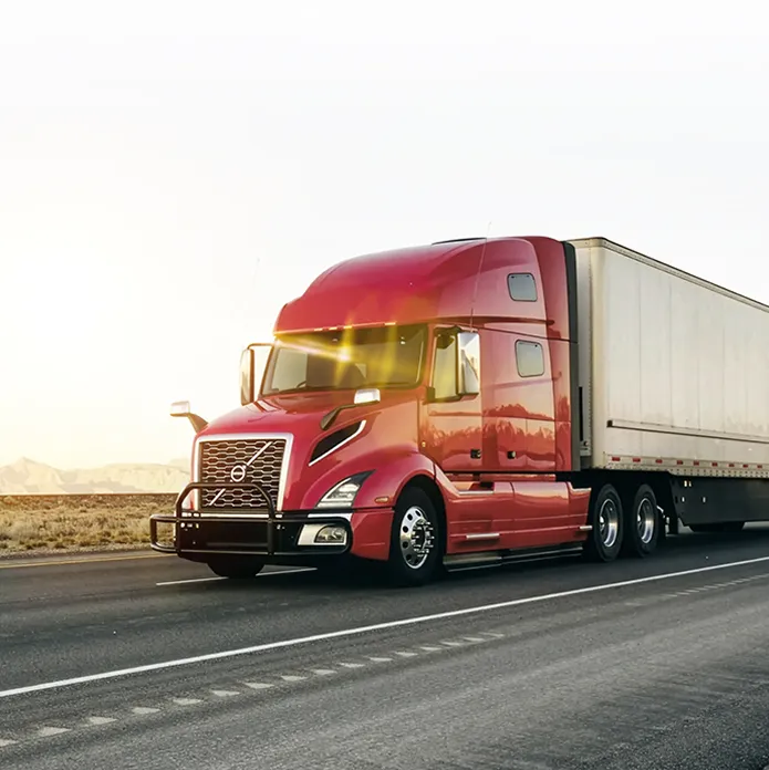 Red semi-truck with large white trailer driving on highway at sunset with desert landscape in background.