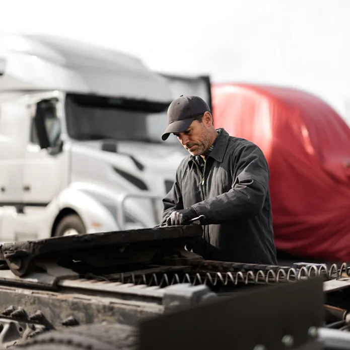 Mechanic in a dark jacket and cap inspecting the coupling area of a truck tractor in front of large trucks.