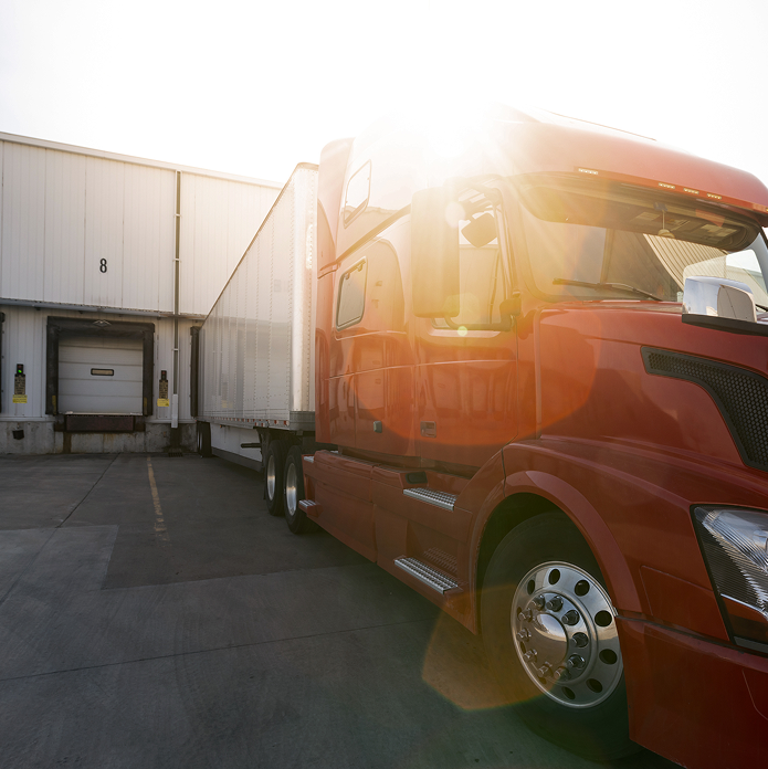 Red semi-truck backed up to a loading dock with sun glare in the background.