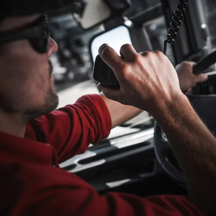 Man wearing sunglasses and red shirt speaking into a handheld radio while driving a vehicle.