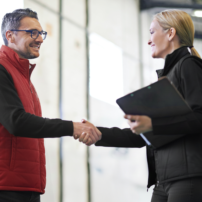Man and woman smiling and shaking hands indoors, woman holding a clipboard.