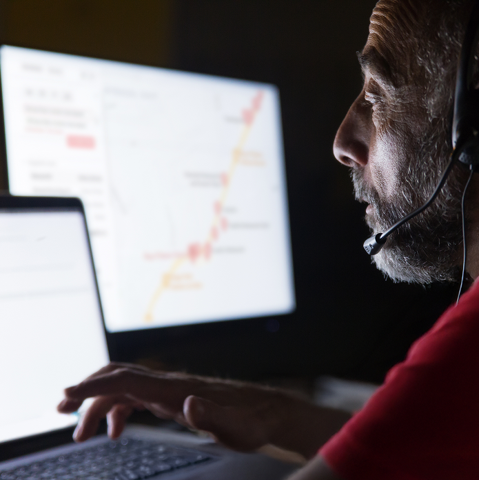 Man wearing a headset working on a laptop with a map displayed on a monitor in a dark room.
