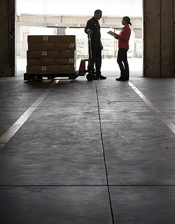Two warehouse workers conversing next to stacked boxes on a pallet jack inside a loading dock.