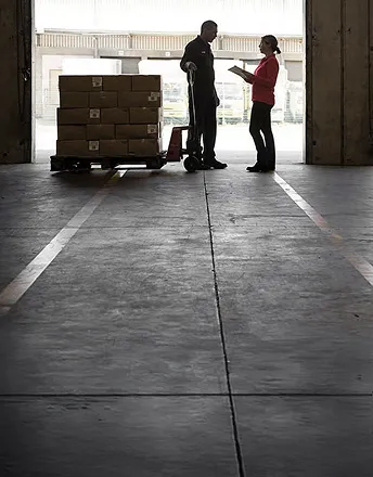 Two warehouse workers conversing next to stacked boxes on a pallet jack inside a loading dock.