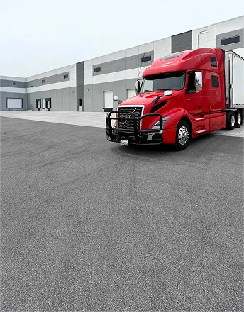 Red semi truck parked outside a modern warehouse building under an overcast sky.
