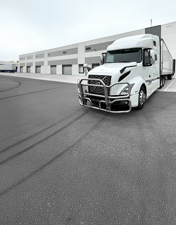 White semi truck with a protective front grille parked near a loading dock at a warehouse.