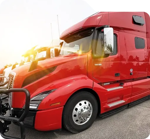 Red semi-truck parked alongside other trucks under a clear sky with sunlight flare.
