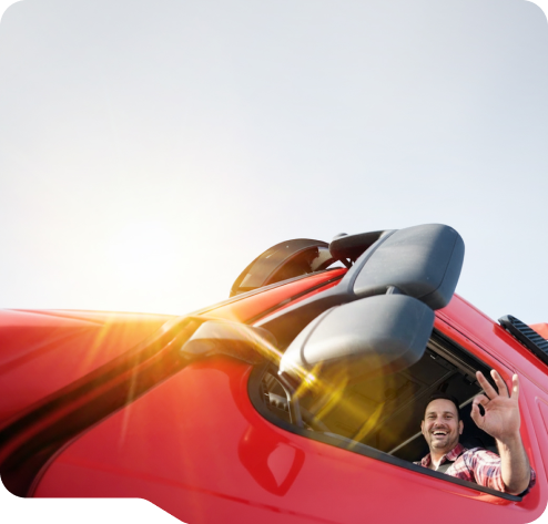 Smiling man waving from the driver's seat of a red truck with sunlight shining through the window.
