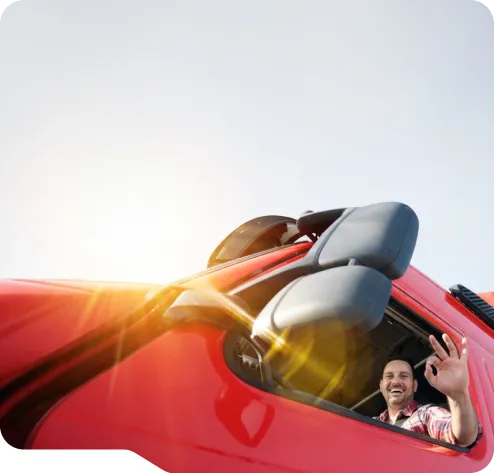 Smiling man waving from the driver's seat of a red truck with sunlight shining through the window.