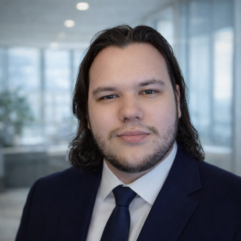 Young man with medium-length dark hair and beard wearing a navy suit, white shirt, and dark tie, posed in an office with blurred windows.