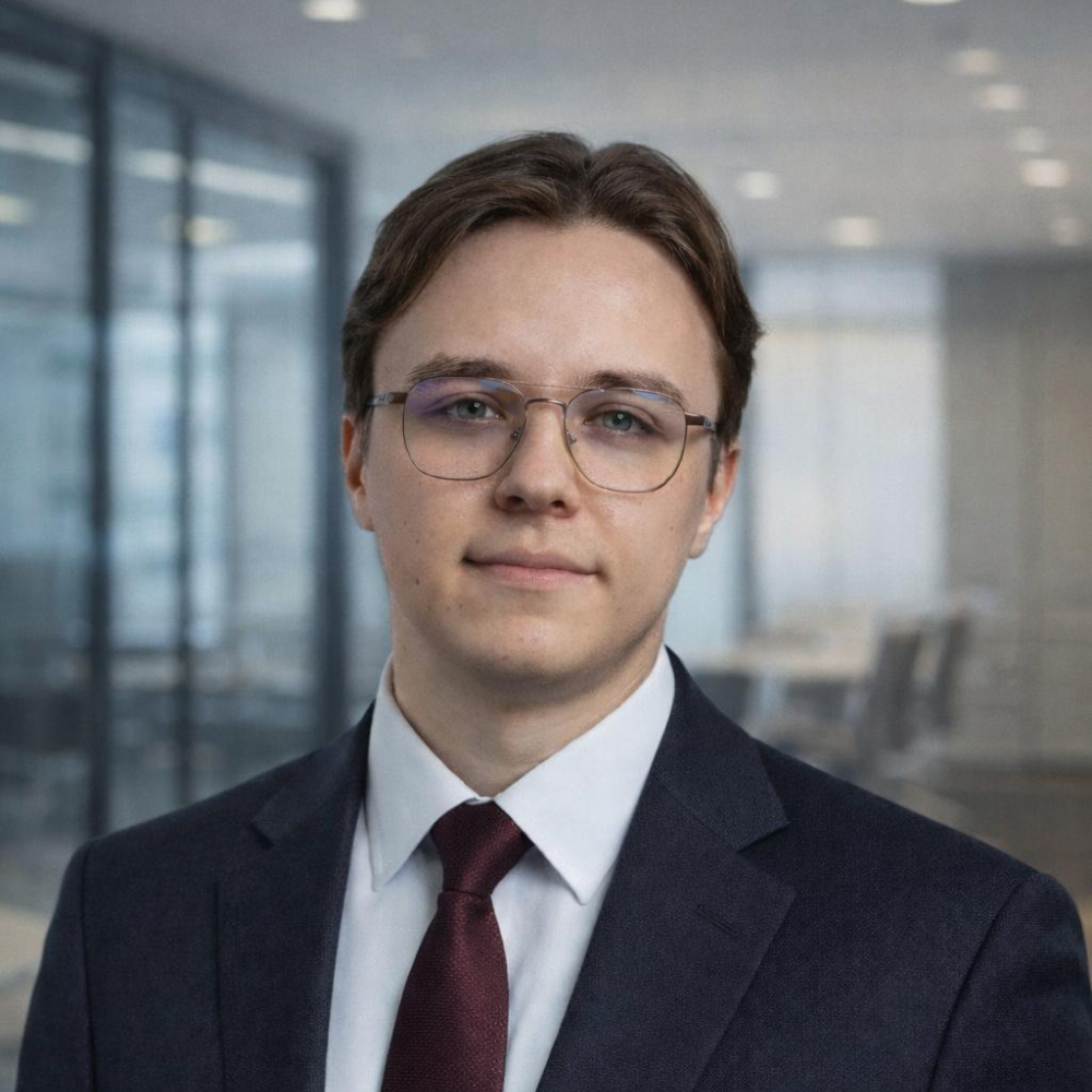 Young man wearing glasses, a dark suit, white shirt, and maroon tie, standing in a modern office environment.