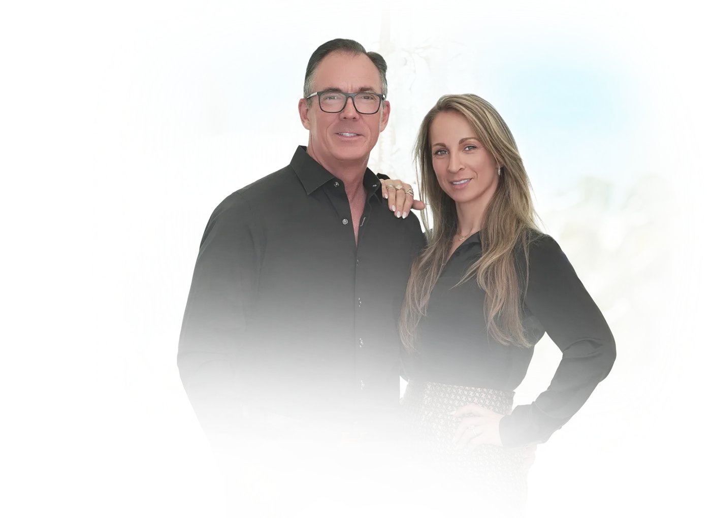 Man and woman standing together, both wearing black shirts, woman with long hair and patterned skirt, man wearing glasses.
