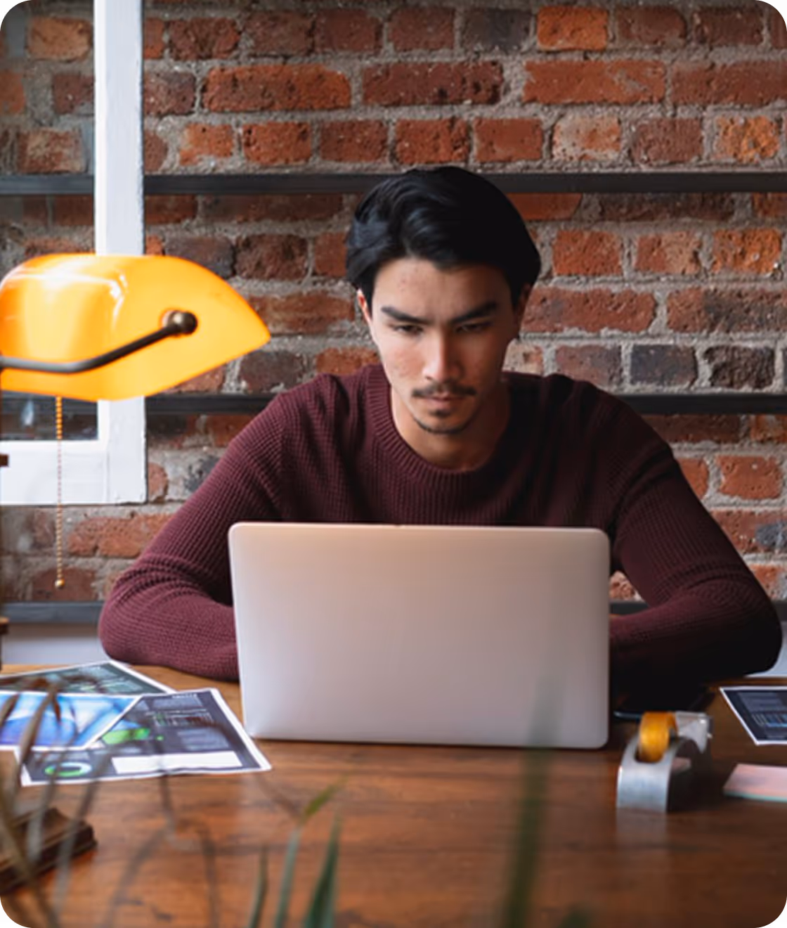 Man in a maroon sweater working on a laptop at a wooden desk with a yellow desk lamp and brick wall background.
