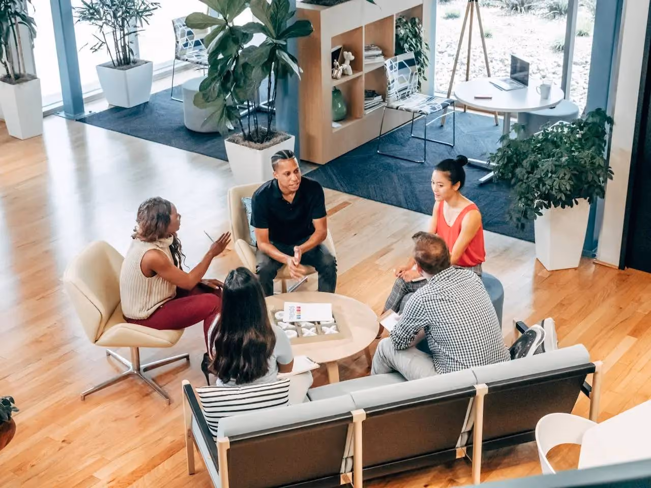 Five diverse colleagues sitting and discussing around a small round table in a modern office lounge with plants and wooden flooring.