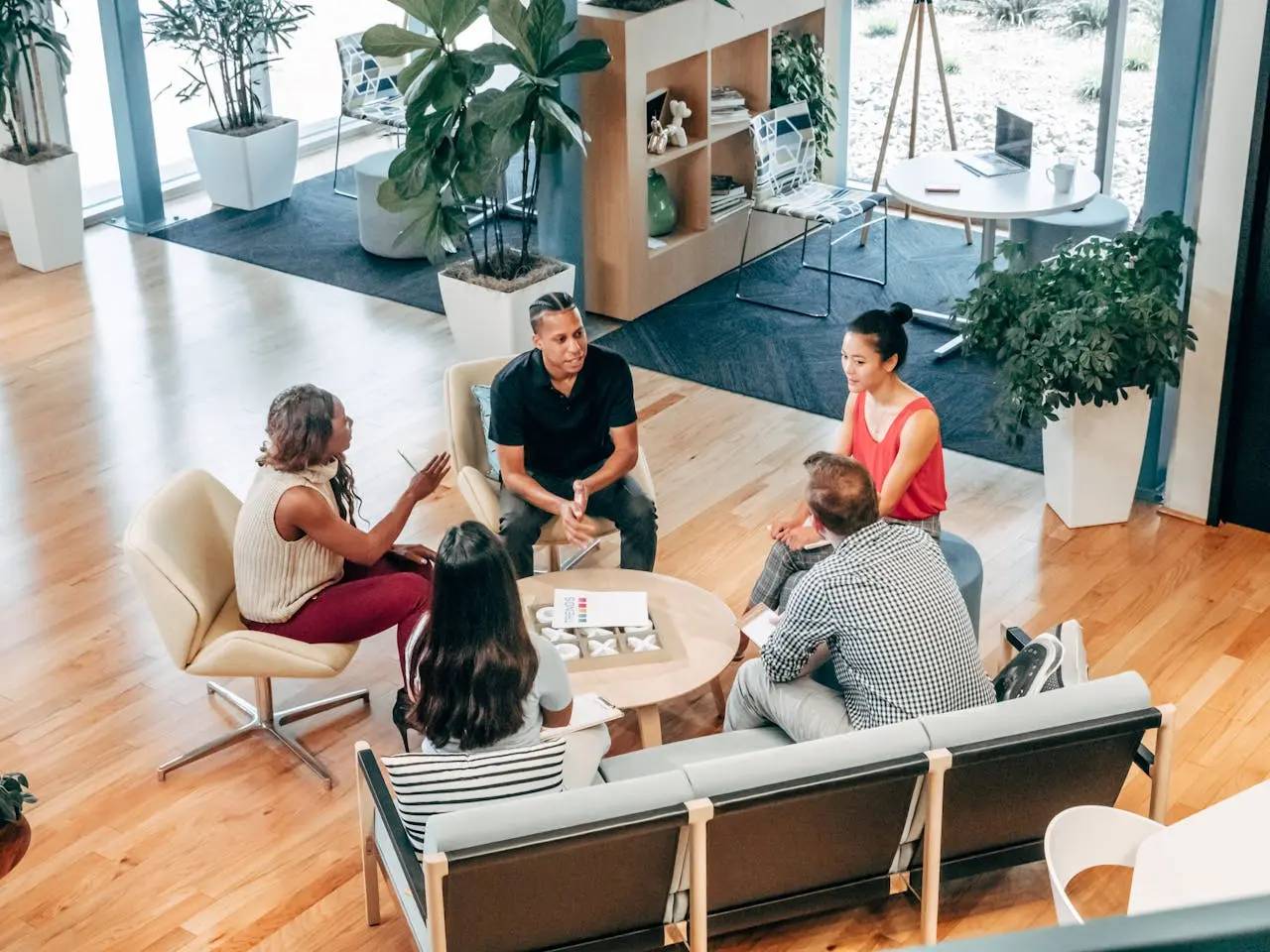 Five diverse colleagues sitting and discussing around a small round table in a modern office lounge with plants and wooden flooring.