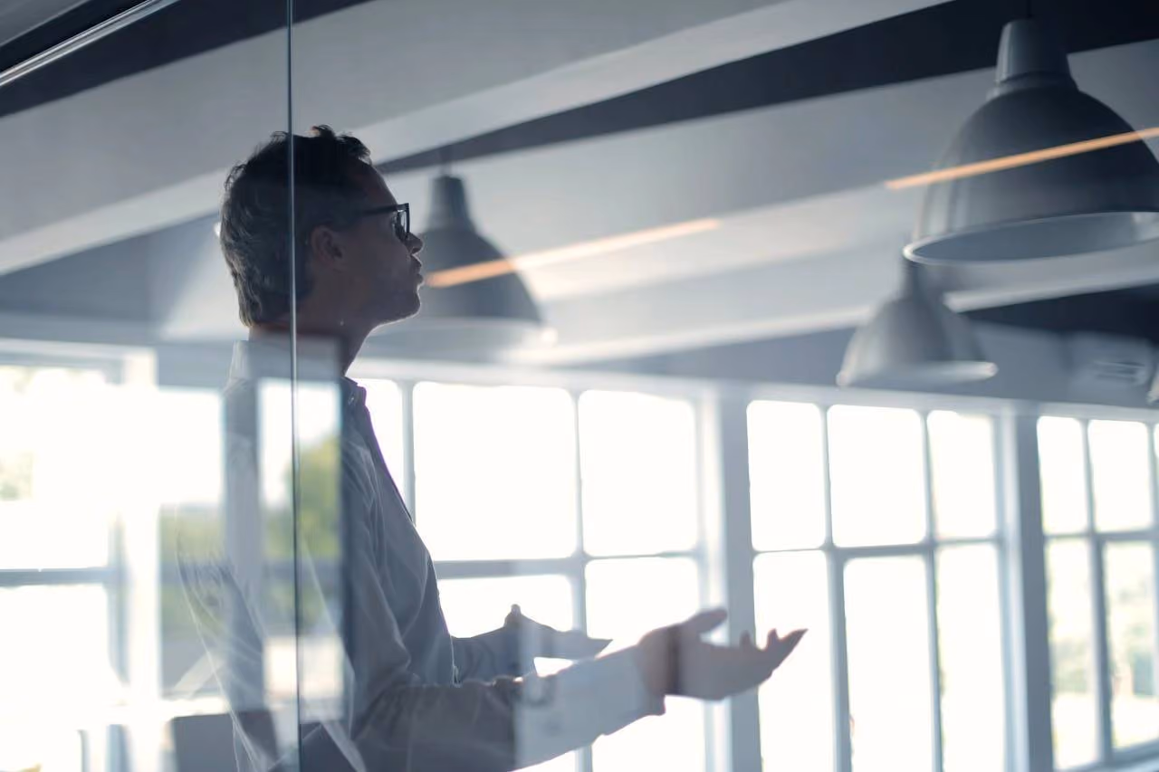 Man wearing glasses gesturing with his hands in an office with large windows and hanging lights.
