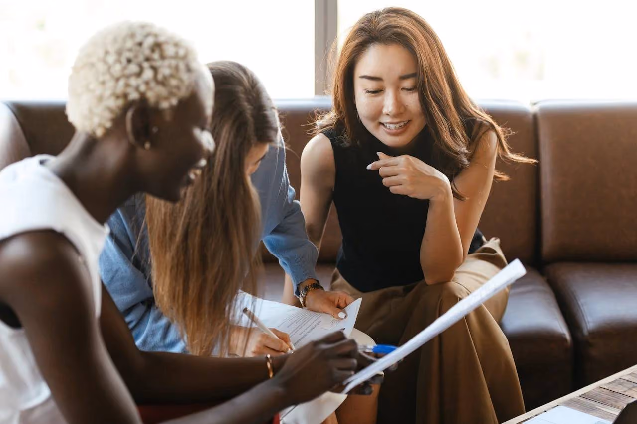 Three women sitting on a couch engaged in reviewing and signing documents together.