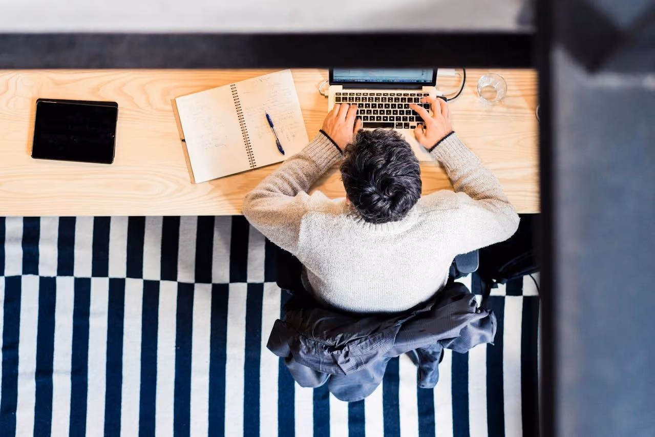 Person typing on a laptop at a wooden desk with an open notebook and a tablet nearby, sitting on a striped black and white rug.