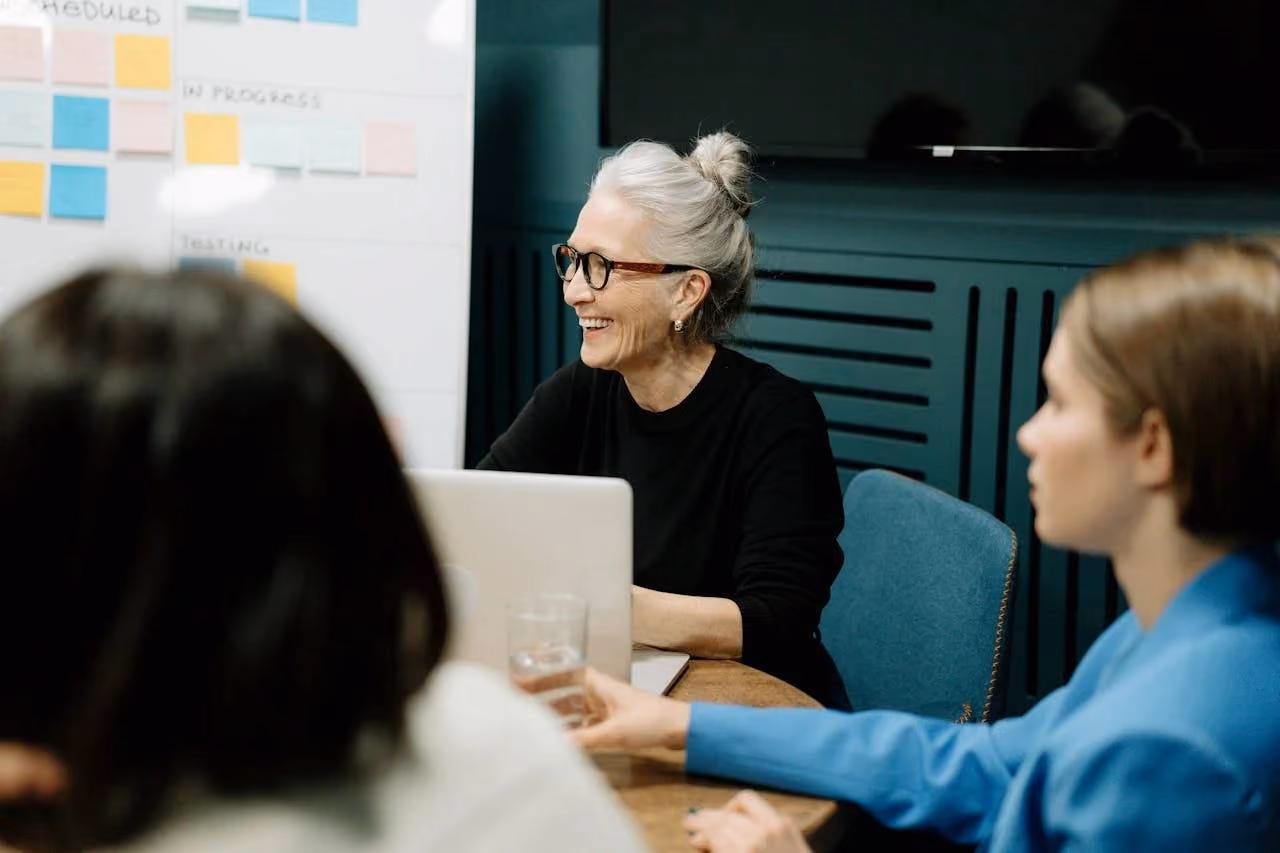 An older woman with gray hair in a bun and glasses smiling while sitting at a table with a laptop during a meeting with two other people.