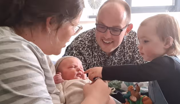 Vader en oudere broer kijken liefdevol naar een pasgeboren baby die door de moeder wordt vastgehouden.