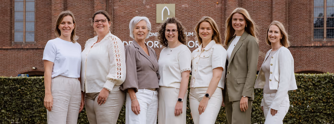 Zeven vrouwen staand in een rij buiten voor een bakstenen gebouw, gekleed in lichte en neutrale kleuren, glimlachend naar de camera.
