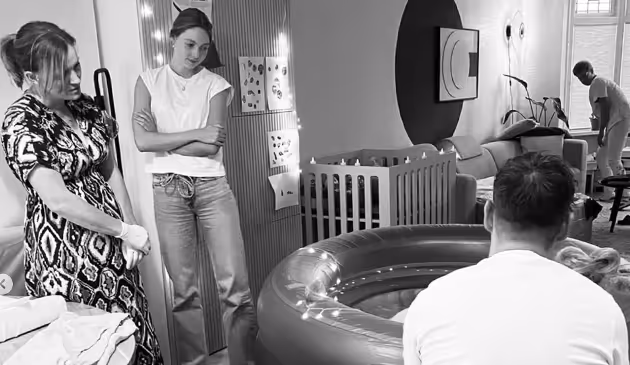 Vier volwassenen in een woonkamer met een luxe geboortebad en een babybedje, voorbereidend op een bevalling.