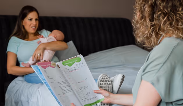 Vrouw die een baby vasthoudt terwijl een andere persoon notities op een bladzijde bekijkt in een slaapkamer.