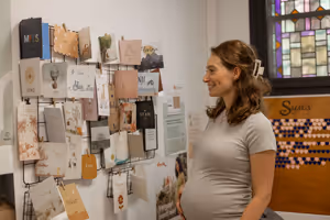 Een zwangere vrouw in een lichtgrijs shirt kijkt glimlachend naar een prikbord met wenskaarten en notities aan een witte muur.