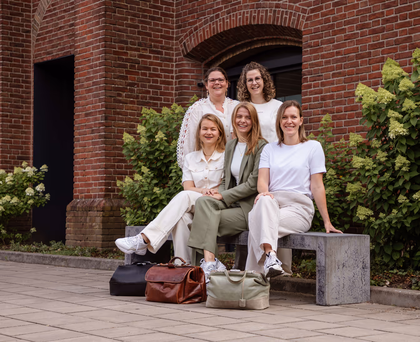 Vijf vrouwen zitten en staan lachend op een stenen bankje buiten, met bruine bakstenen muur en groene planten op de achtergrond.