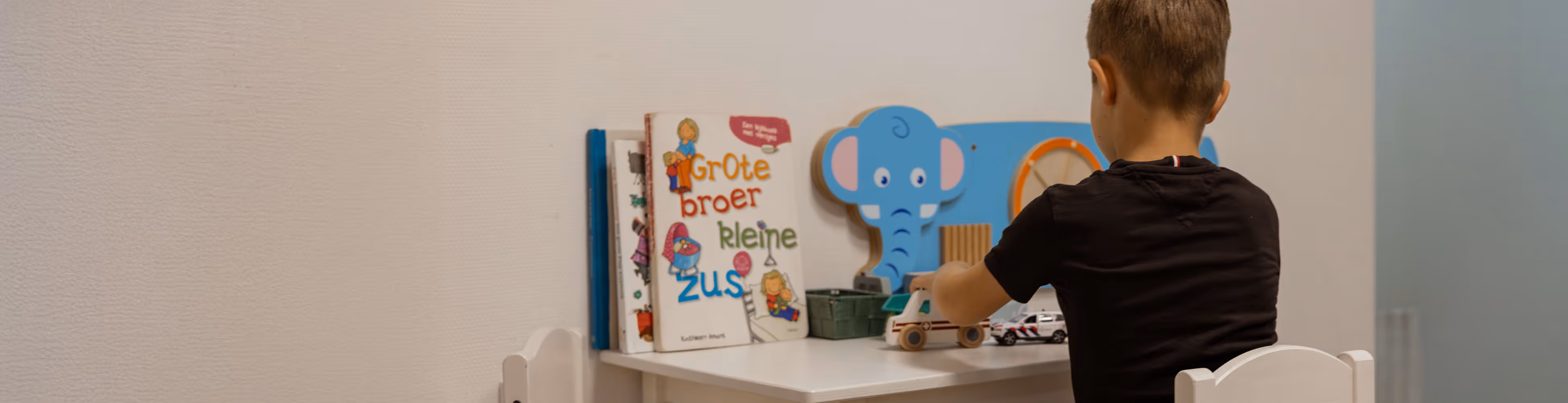 Een jongen zit aan een witte tafel en speelt met speelgoedauto's, met een stapel kinderboeken en een blauwe olifant speelgoed aan de muur.
