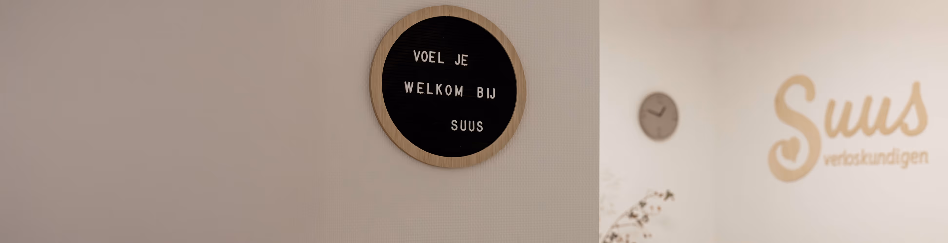 Ronde zwarte tekstbord met houten rand aan een lichtgekleurde muur met de tekst: 'Voel je welkom bij Suus', naast een muur met het woord 'Suus verloskundigen' en een klok.