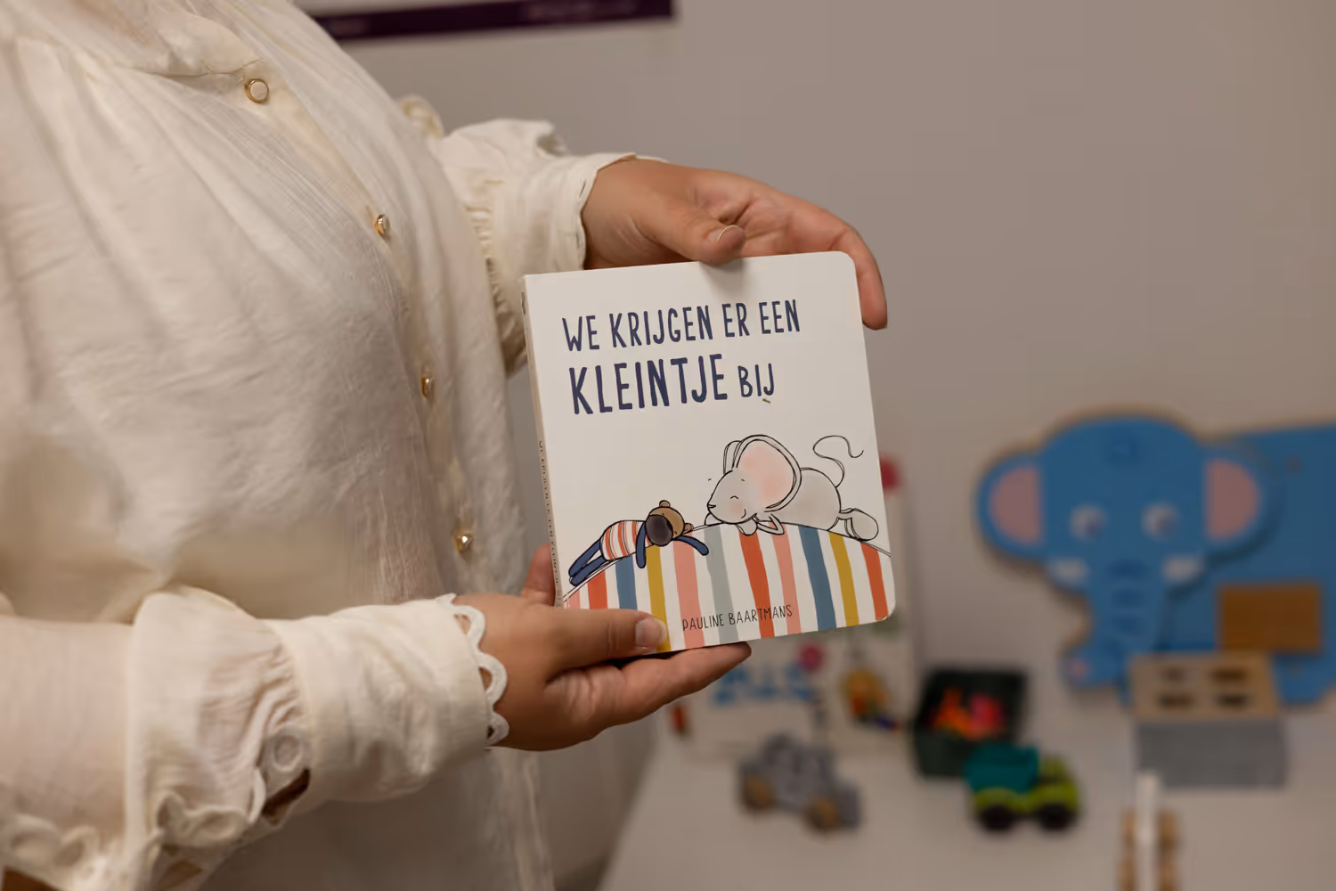 Persoon houdt kinderboek 'We krijgen er een kleintje bij' met illustratie van een muis en een beer op een gestreepte heuvel.