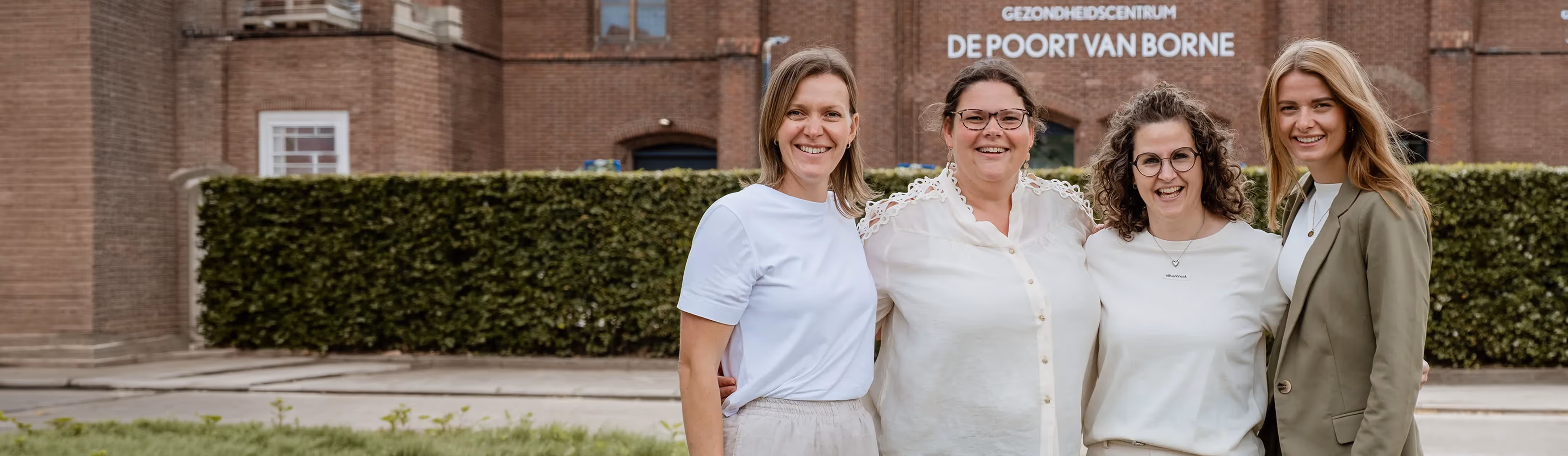 Vier vrouwen die glimlachen, staand voor een gebouw met het bord Gezondheidscentrum De Poort van Borne.