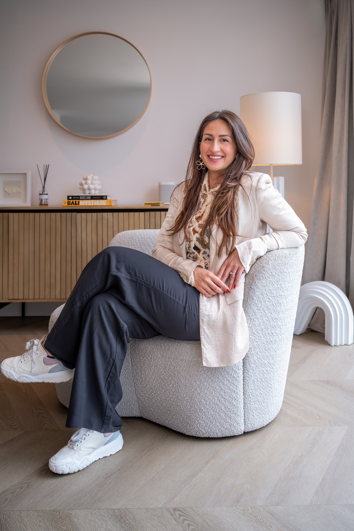 Vrouw met lang haar, beige blazer en zwarte broek zit ontspannen op een moderne, zachte fauteuil in een stijlvolle woonkamer.