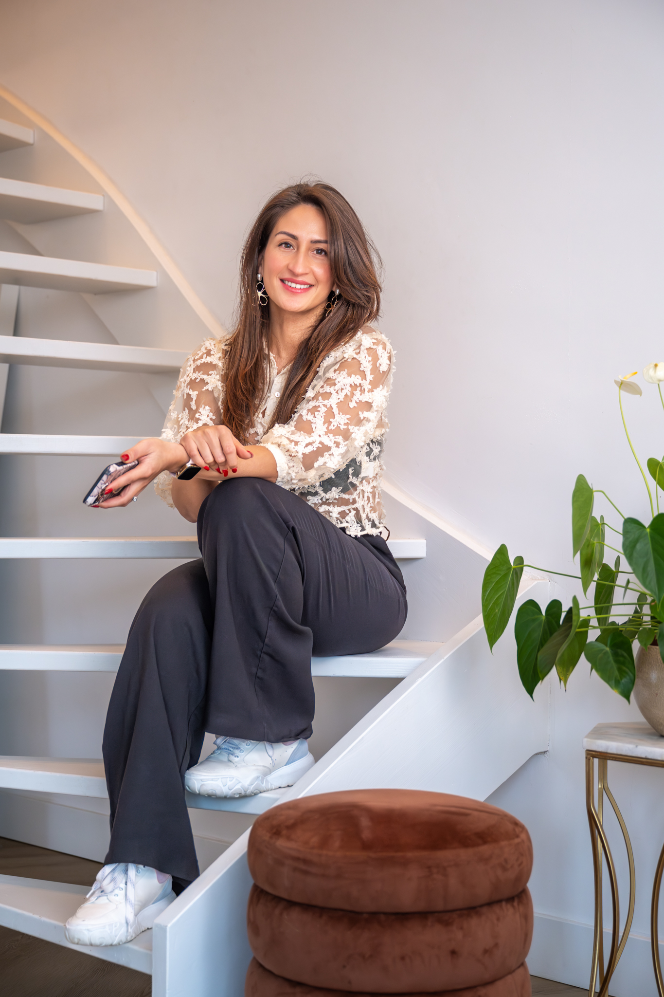 Vrouw met lang haar en doorzichtige kanten blouse zittend op een witte trap, naast een groen bladplant en een bruine poef.