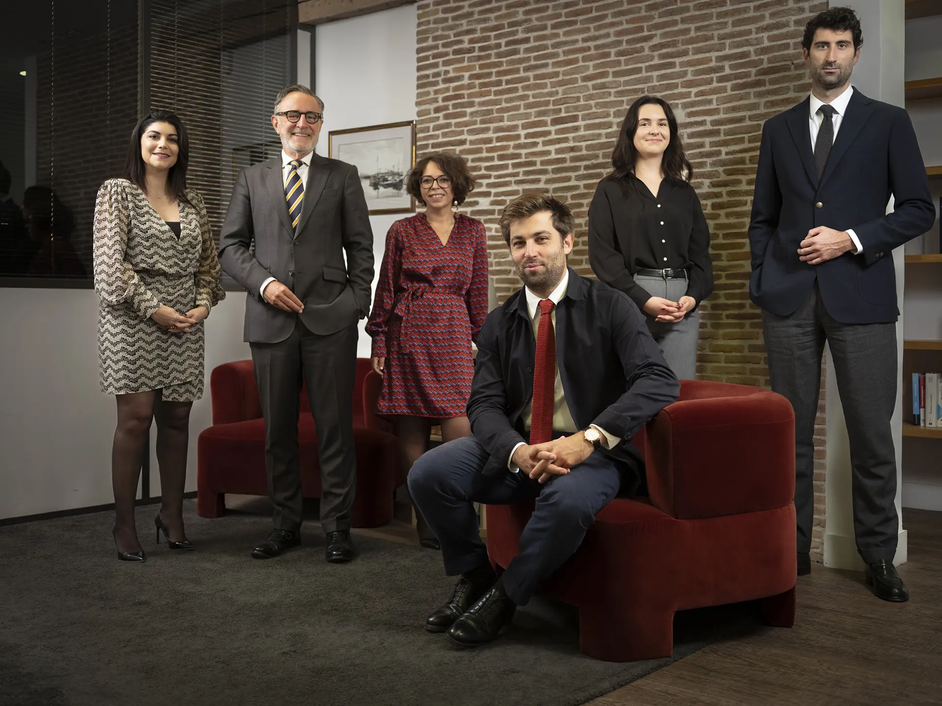 Groupe de six professionnels souriants, cinq debout et un assis sur un fauteuil rouge, dans un bureau moderne avec un mur en briques.
