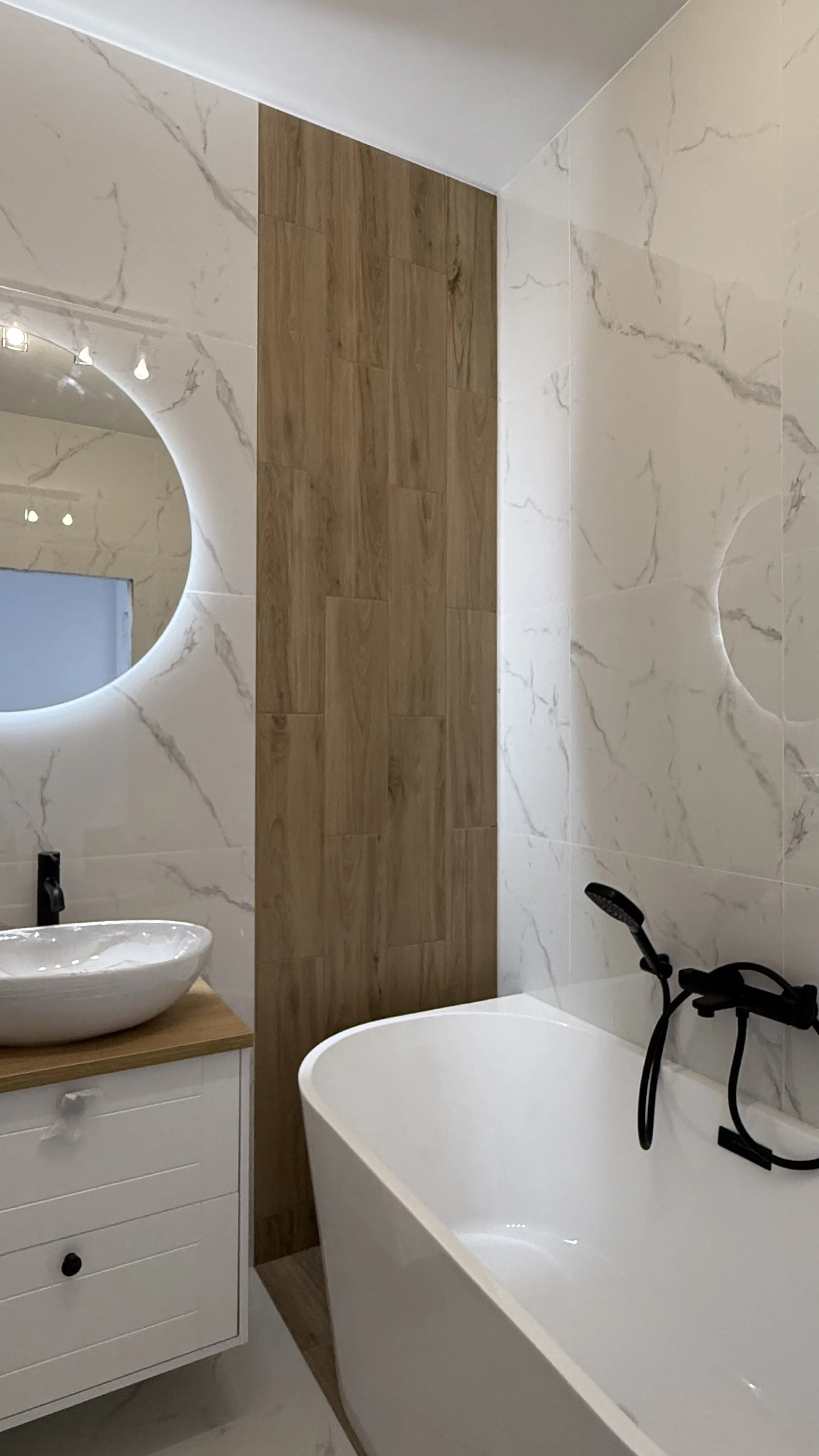 Nowoczesna łazienka z białą wolnostojącą wanną, czarną baterią i umywalką na drewnianym blacie oraz oświetlonym okrągłym lustrem na tle płytek imitujących marmur i drewno.