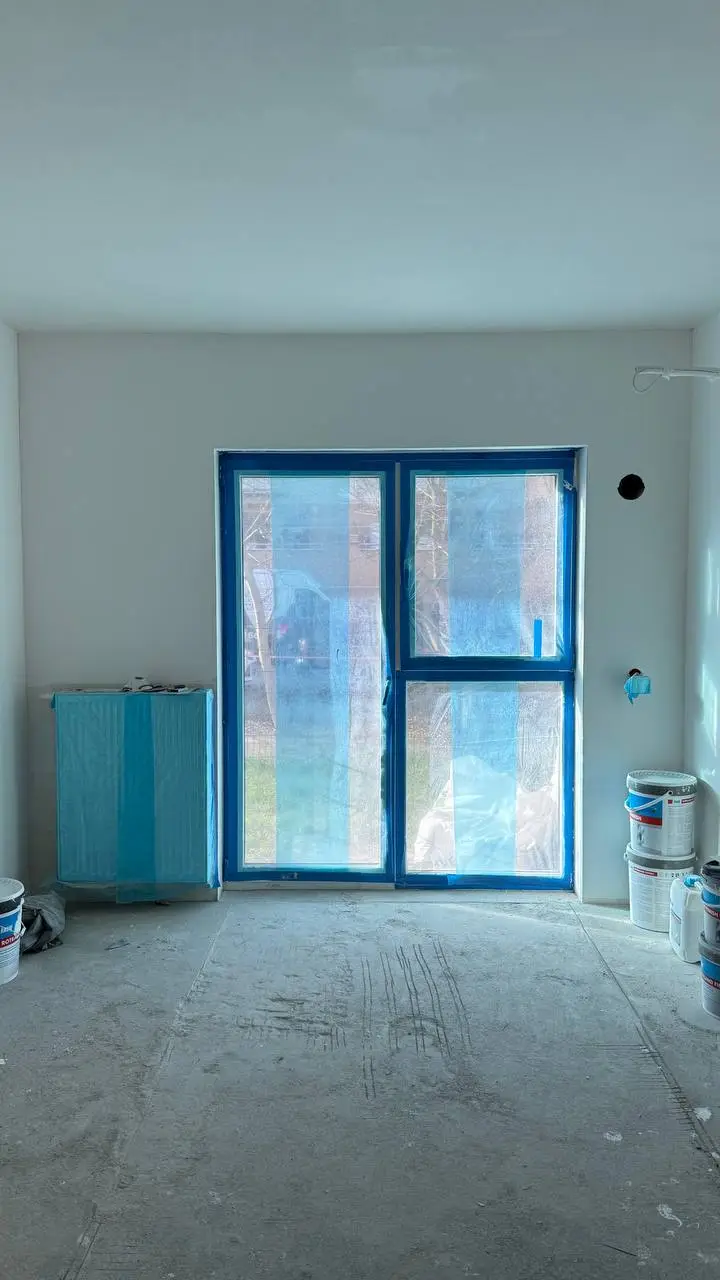 Puste pomieszczenie w trakcie remontu z oknem i drzwiami balkonowymi zabezpieczonymi folią ochronną oraz farbami i innymi materiałami malarskimi na podłodze i przy ścianach.