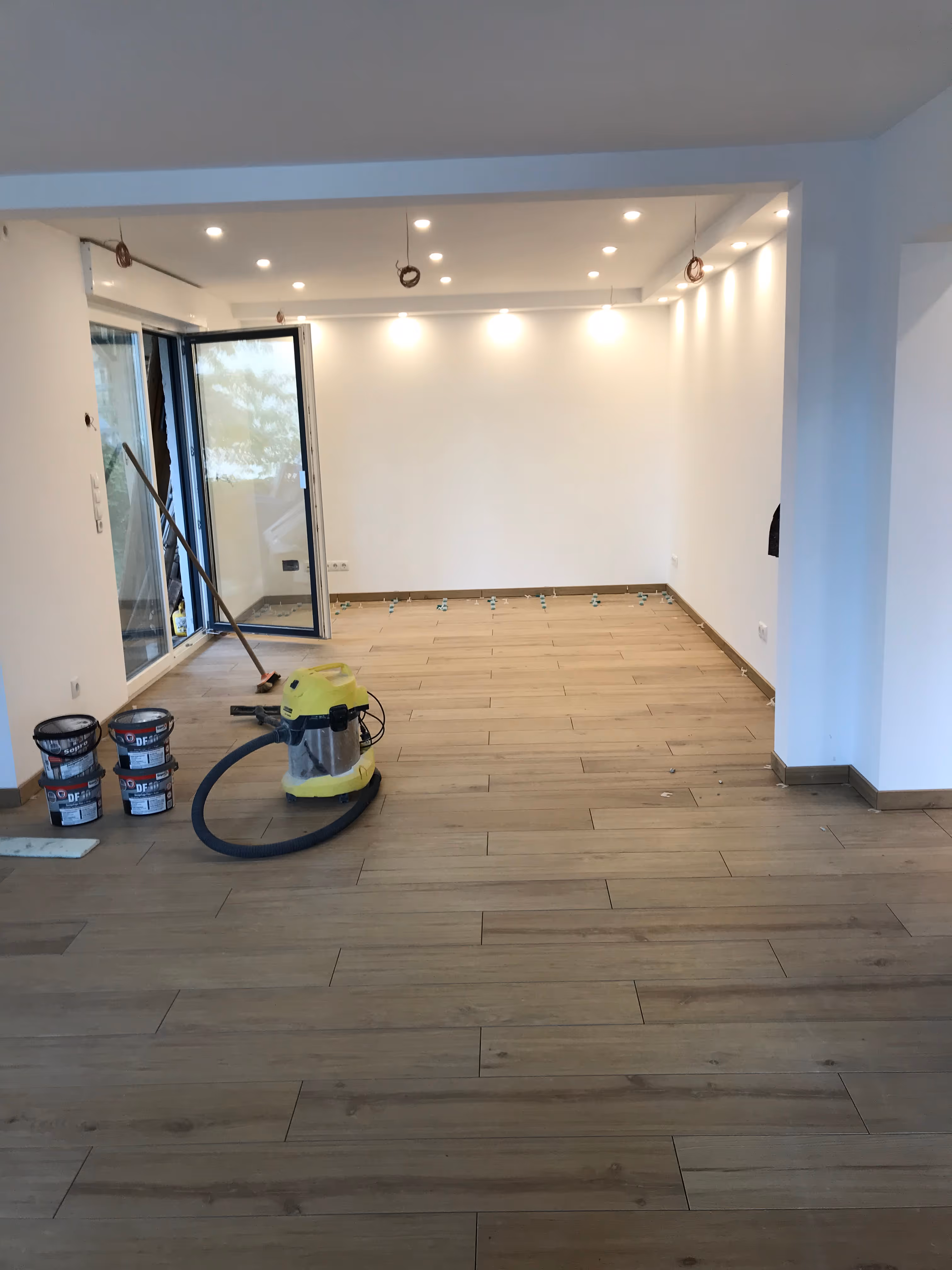 Puste pomieszczenie z niedokończonym montażem podłogi z drewnianych paneli i wyposażone w otwarte drzwi balkonowe oraz narzędzia budowlane.