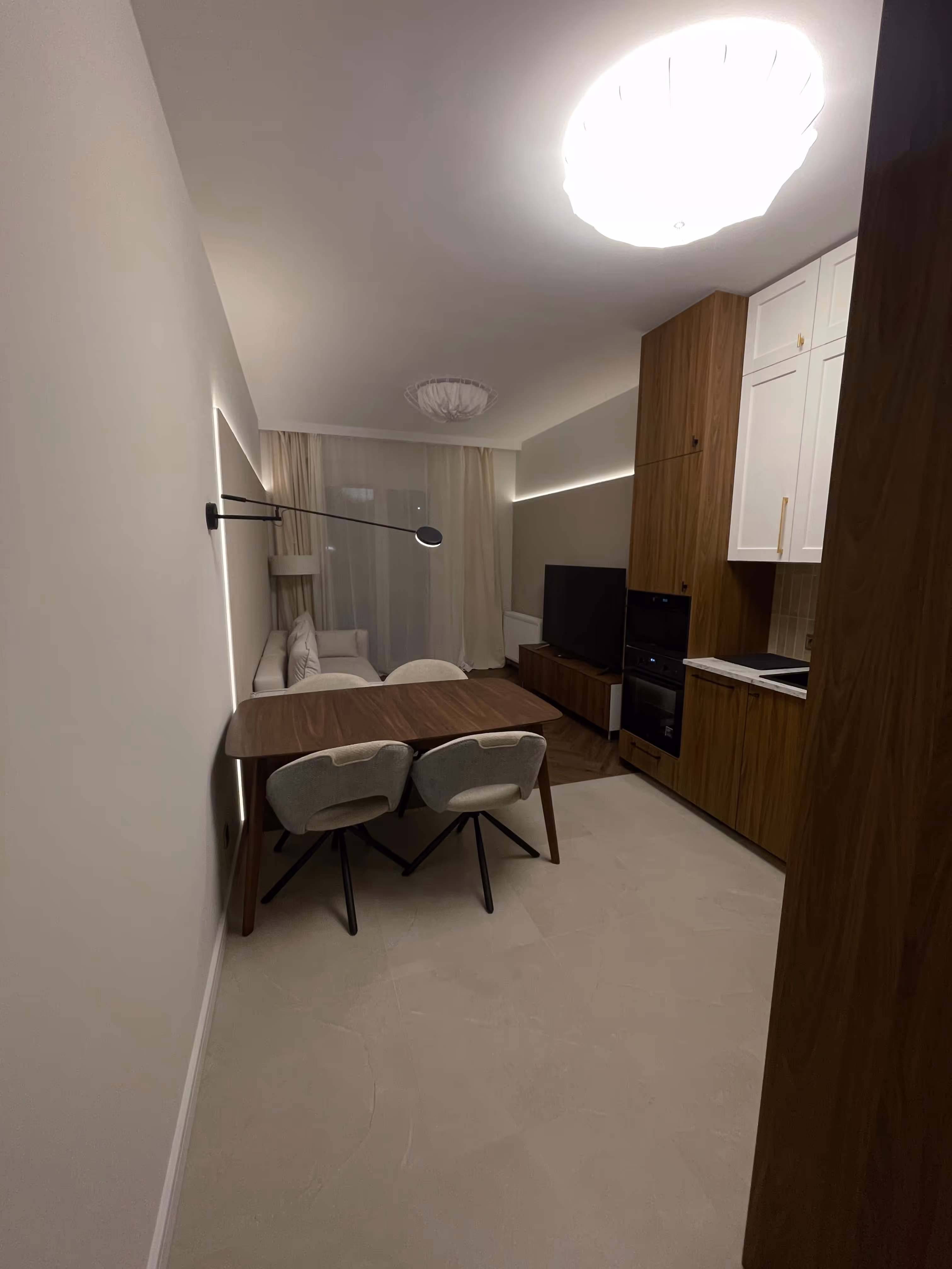 Nowoczesne mieszkanie z aneksem kuchennym, drewnianym stołem, dwoma szarymi krzesłami, białą sofą i dużym telewizorem.