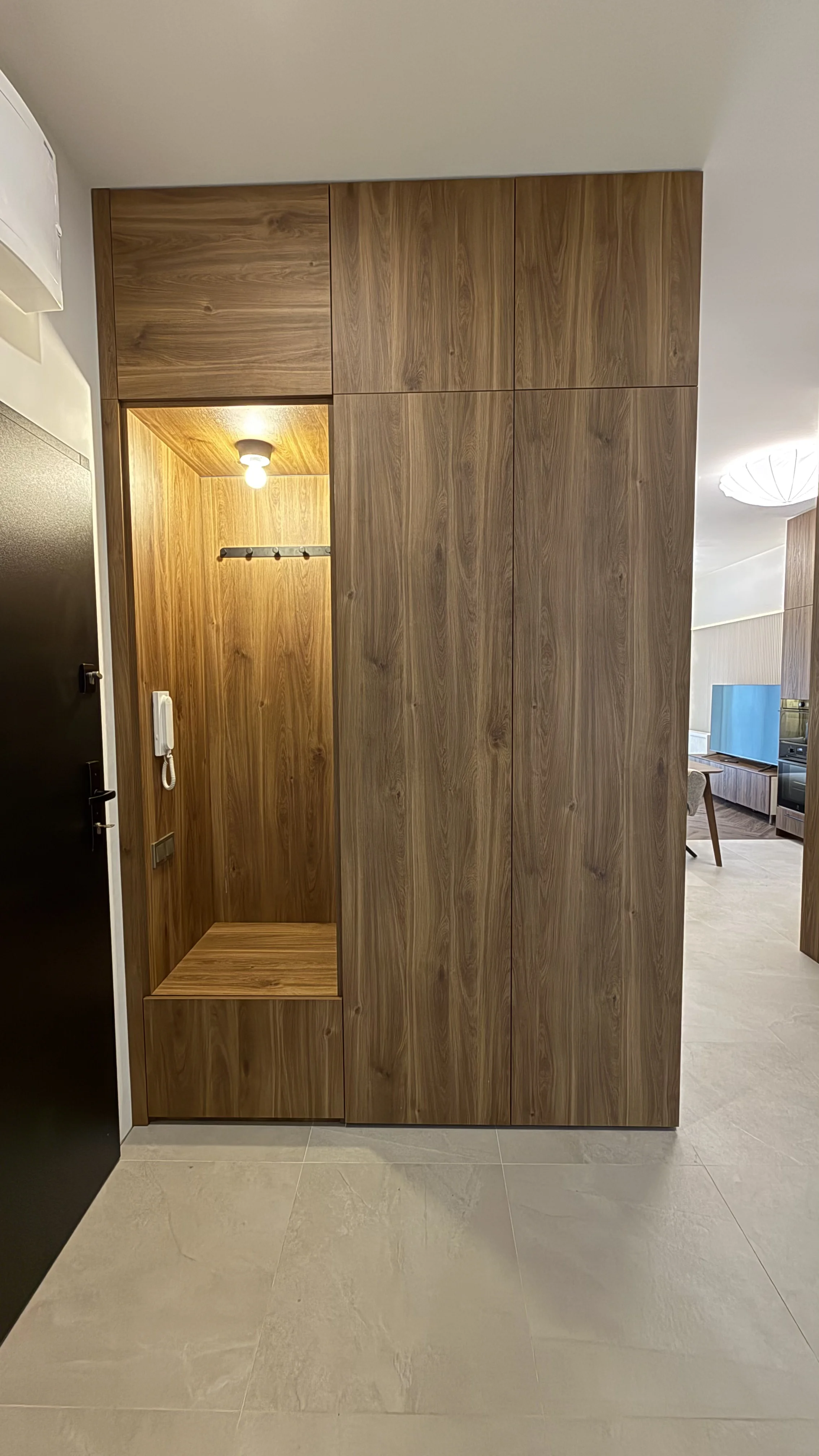 Wąskie wejście z wbudowaną drewnianą szafką na odzież i siedziskiem, oświetlone lampą sufitową, obok drzwi wejściowych.