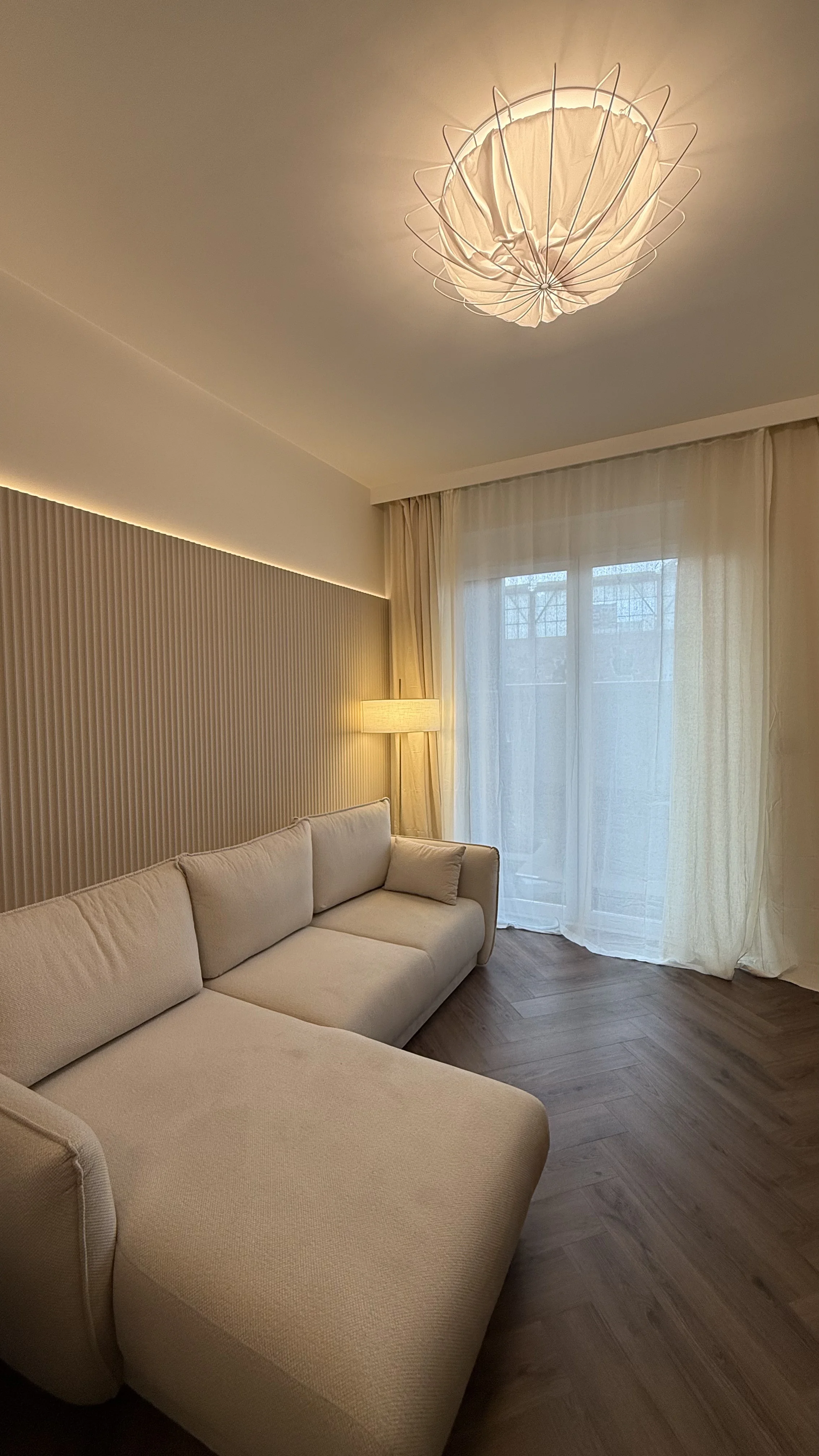 Beżowa sofa w pokoju z drewnianą podłogą, lampą stojącą przy ścianie i dużym oknem z przezroczystymi zasłonami.