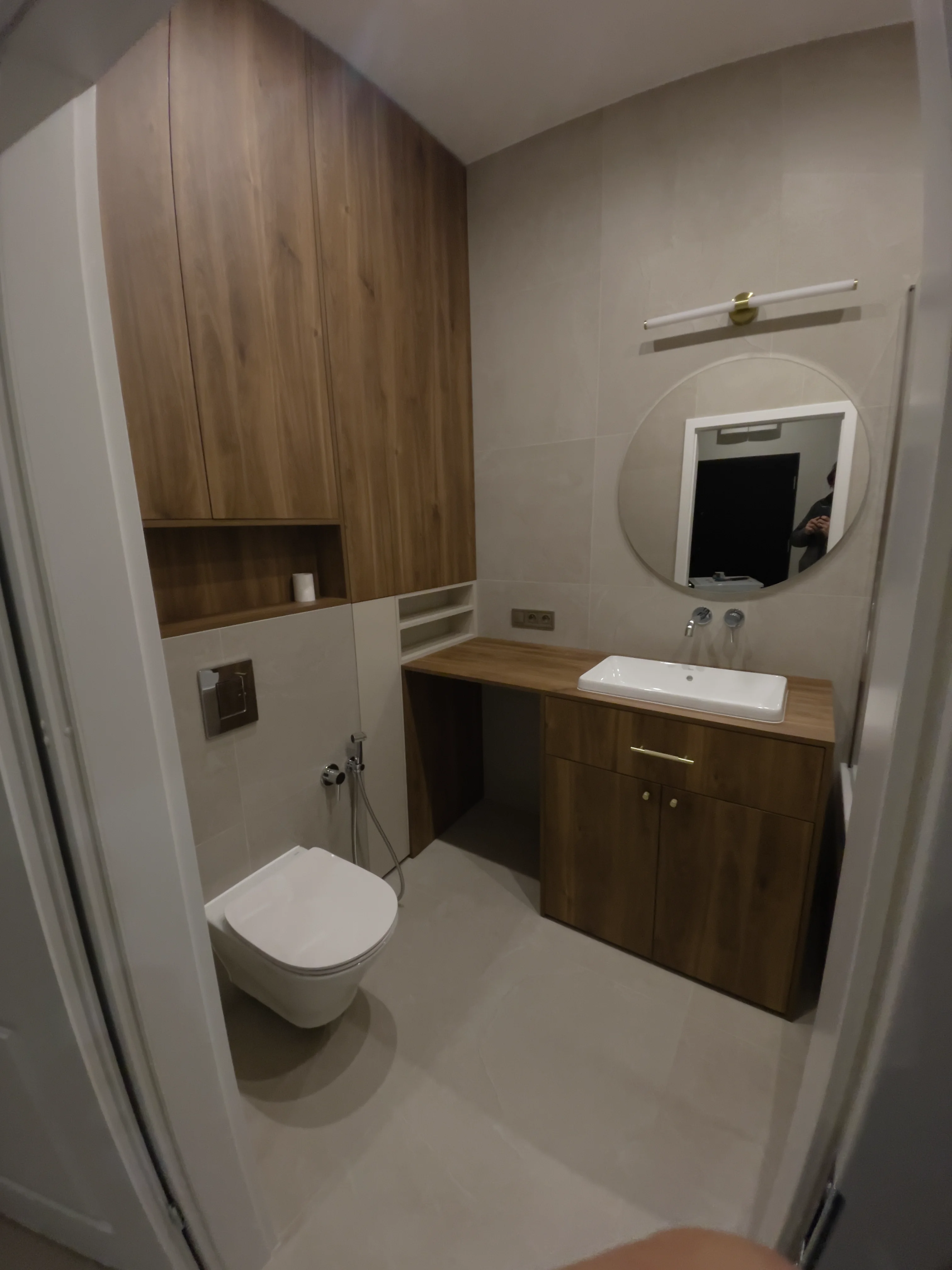 Nowoczesna łazienka z wiszącą muszlą klozetową, drewnianymi szafkami, umywalką i okrągłym lustrem na jasnych płytkach.