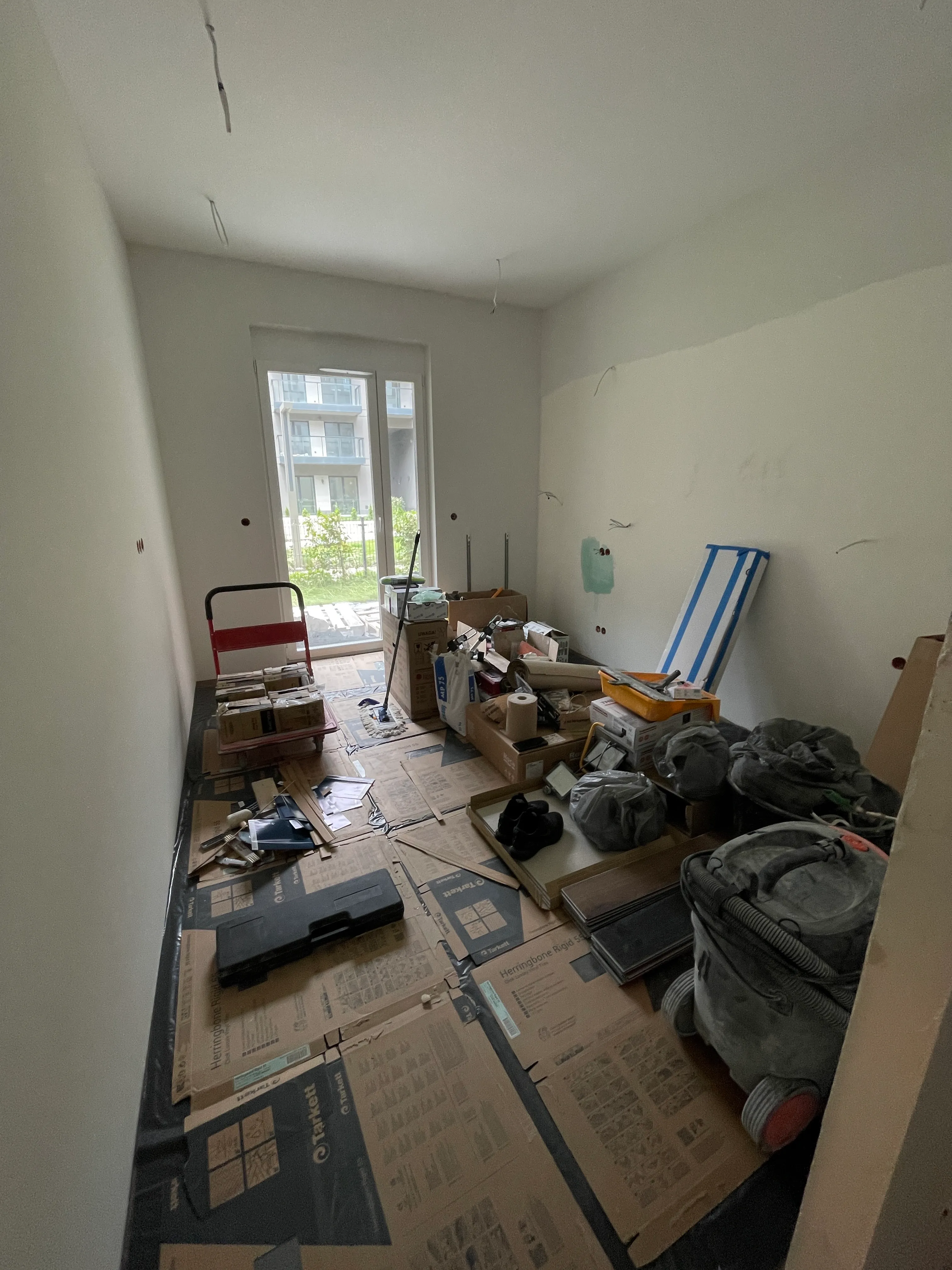 Pokój z kartonami i narzędziami do remontu na podłodze, widok na drzwi balkonowe z ogrodem na zewnątrz.