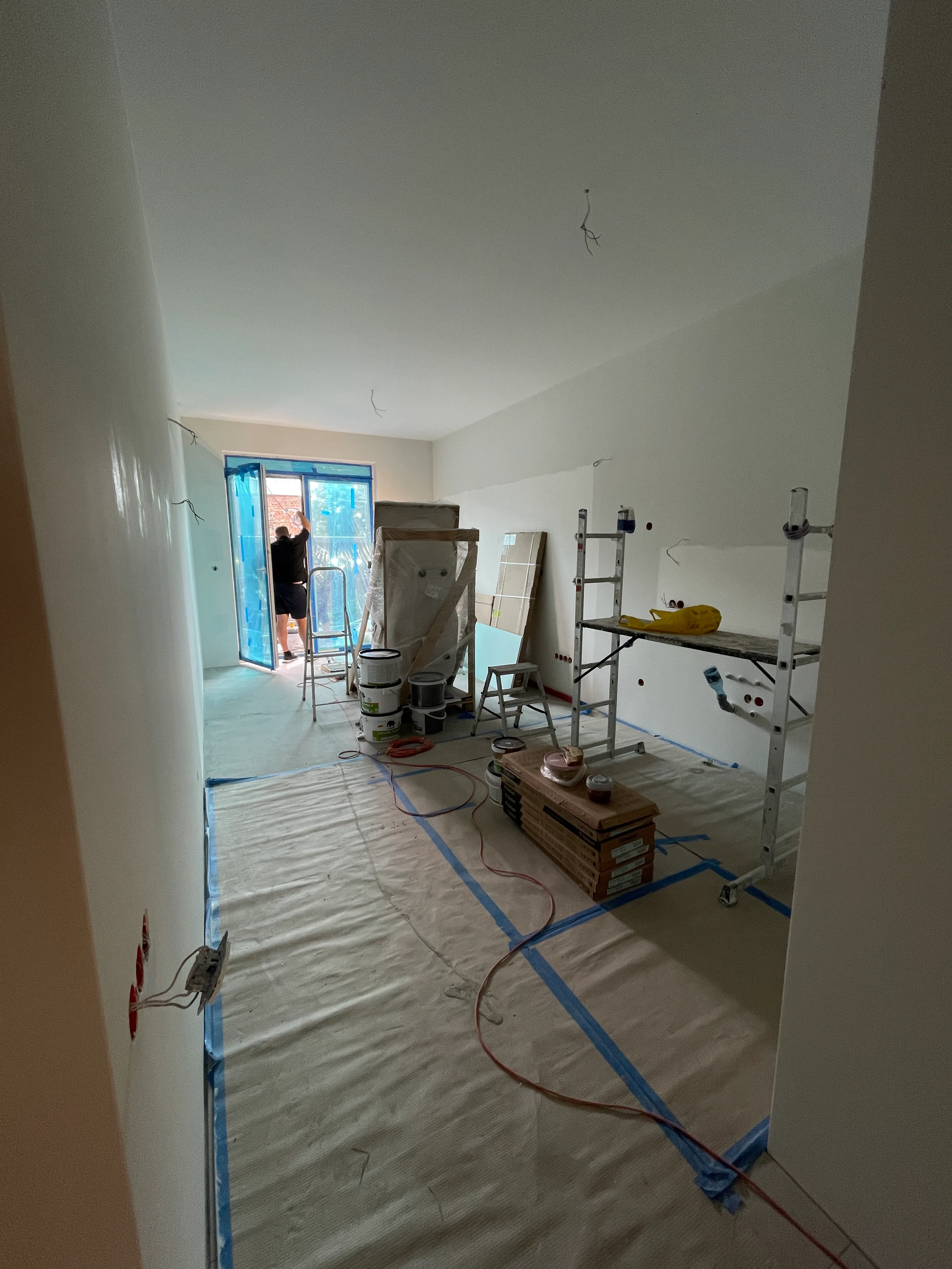 Pomieszczenie podczas remontu z zabezpieczoną podłogą, narzędziami, farbami oraz mężczyzną stojącym przy otwartym drzwiach na balkon.