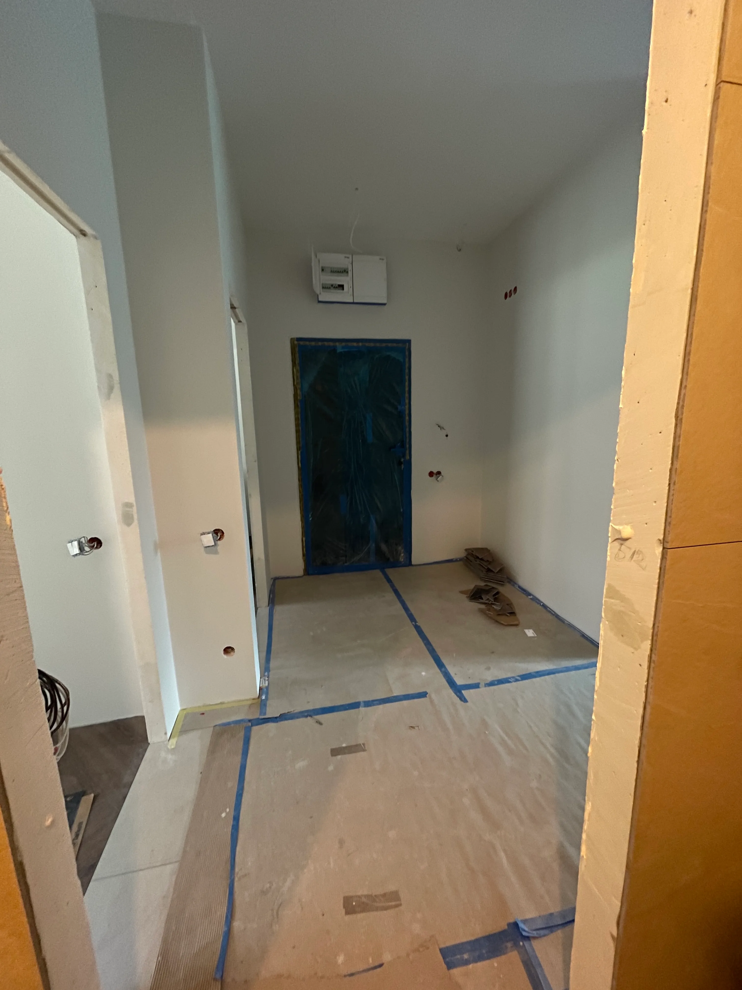 Pokój w trakcie remontu z białymi ścianami, zabezpieczoną taśmą podłogą i drzwiami zakrytymi folią ochronną.