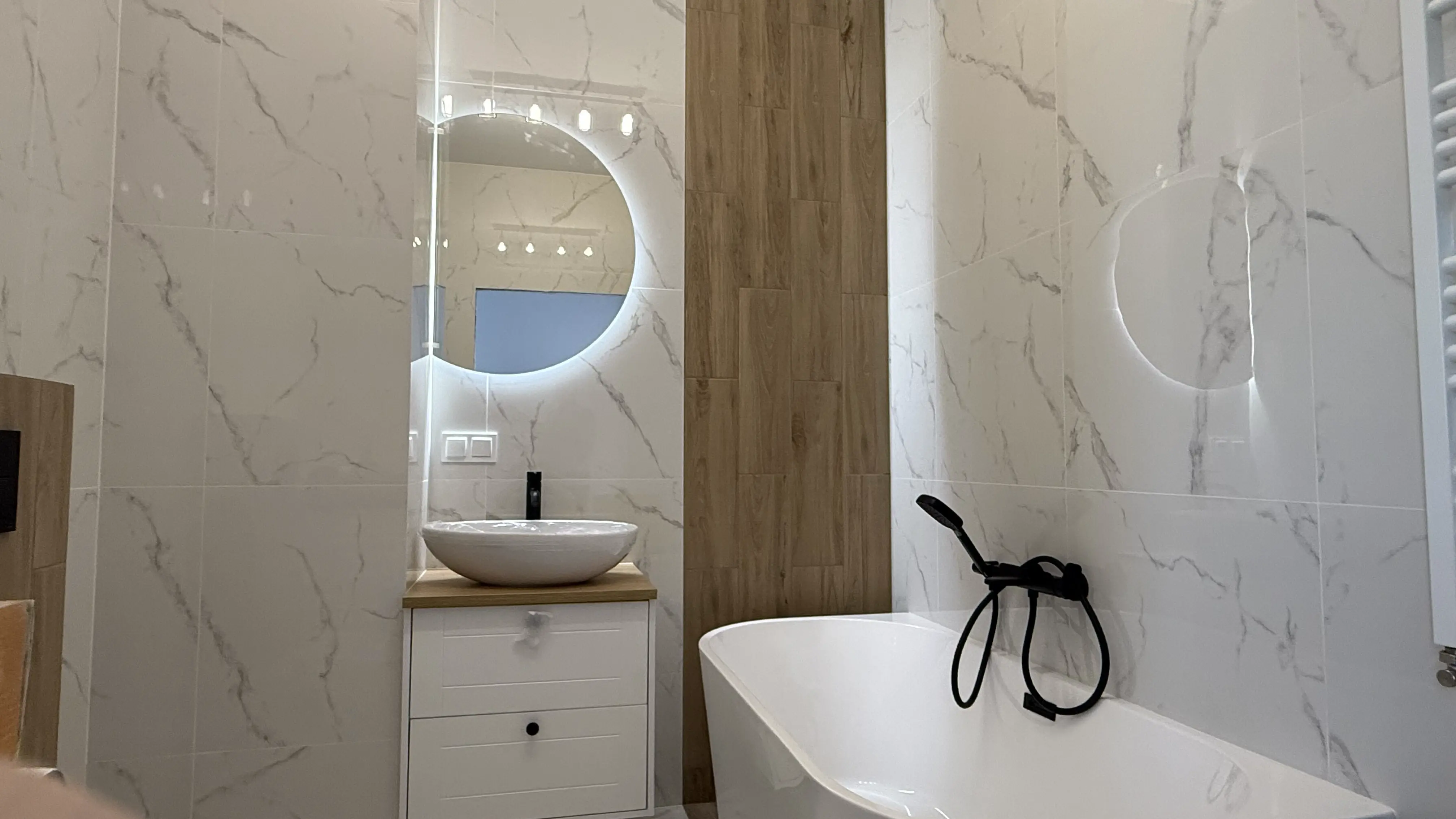 Nowoczesna łazienka z okrągłym podświetlanym lustrem, umywalką na blacie drewnianym oraz białą wanną z czarną baterią.